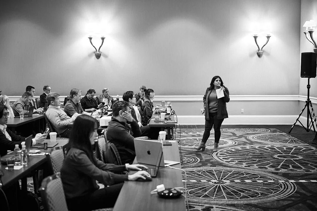 A woman speaking on stage with a microphone to an audience seated at tables, some using laptops, in a conference room with patterned carpet and wall-mounted lights.