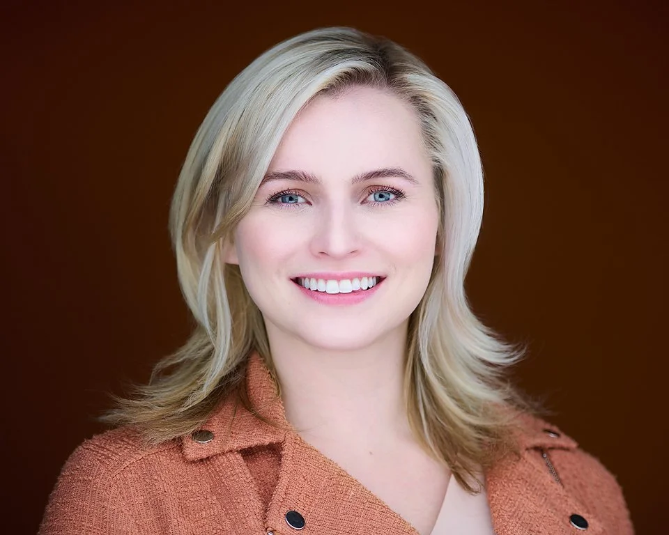 Portrait of a smiling woman with blonde hair, blue eyes, wearing a brownish-orange jacket, against a brown background.