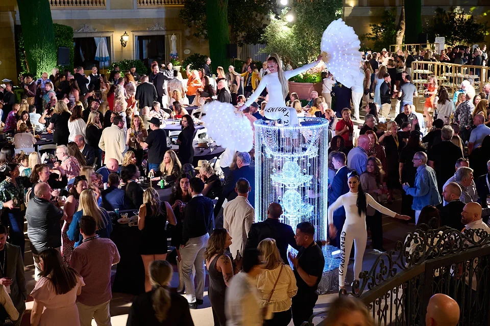 A crowded dance party with people dancing and socializing outdoors at night. A performer on stilts with illuminated white wings and a white costume is in the center, celebrating with the crowd. There are tables with seated guests, greenery, and strin