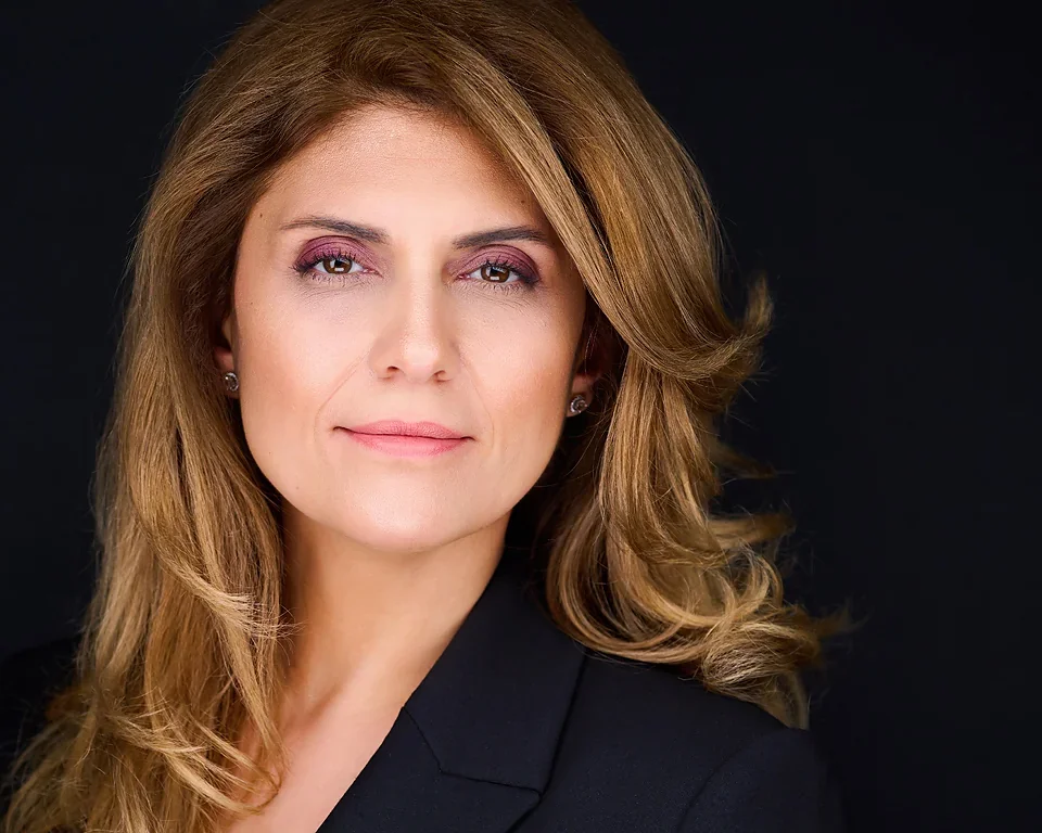 A woman with long, light brown hair and a confident expression, wearing a black blazer against a dark background.