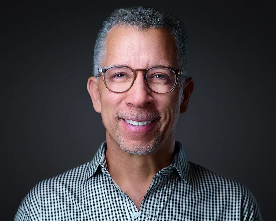Smiling middle-aged man with gray curly hair and glasses, wearing a checkered shirt, against a dark background.