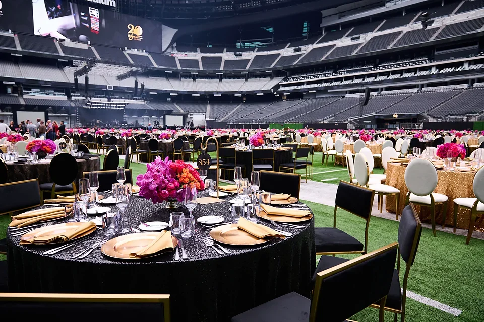 Indoor stadium set up for a formal event with round tables, pink floral centerpieces, and black and white chairs, with a large audience area in the background.
