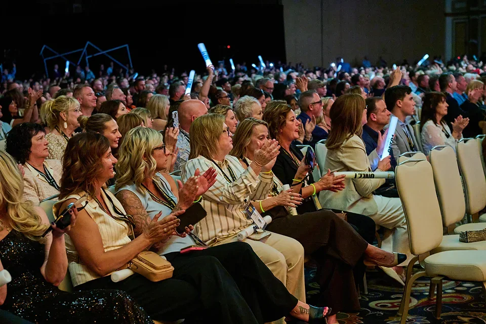 Audience members sitting in rows at a conference or event, some clapping and using glow sticks.