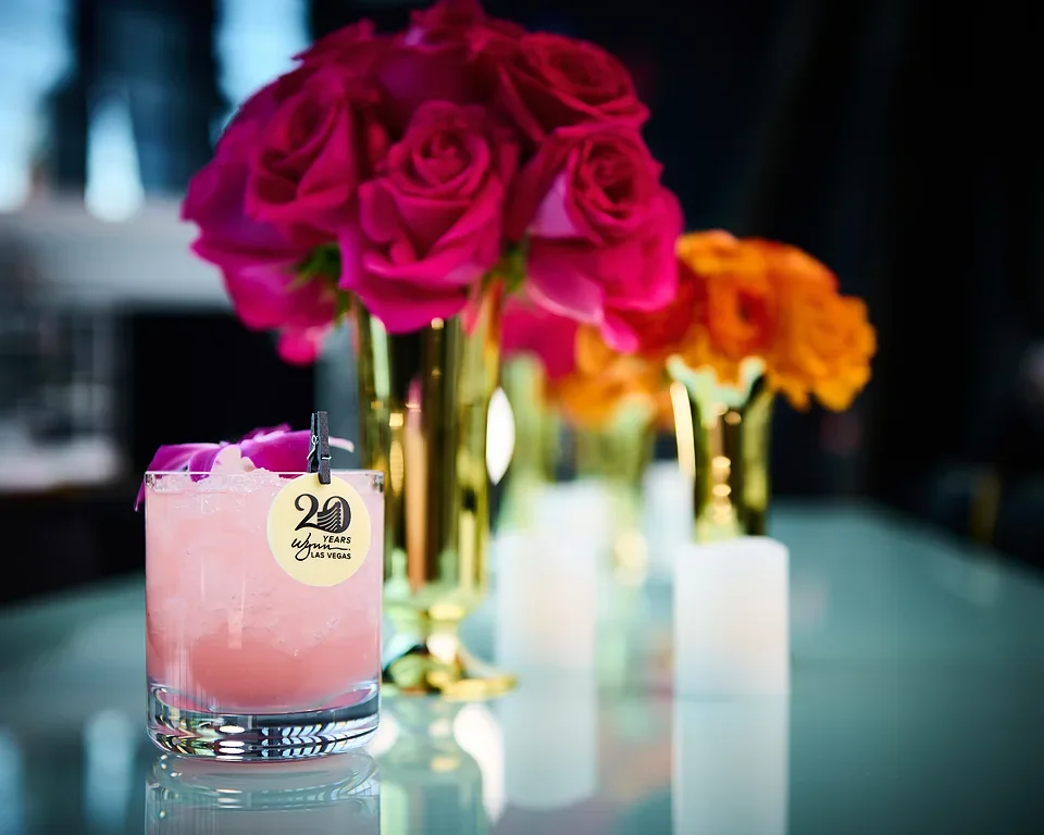 Pink cocktail with ice and a lemon slice garnishment on a glass, with pink and orange flowers in gold vases in the background.
