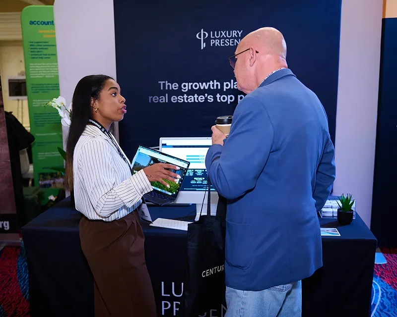 Two professionals engaged in conversation at a business conference booth, with a large banner reading 'Luxury Presentation' and 'The growth plan for real estate's top' in the background.