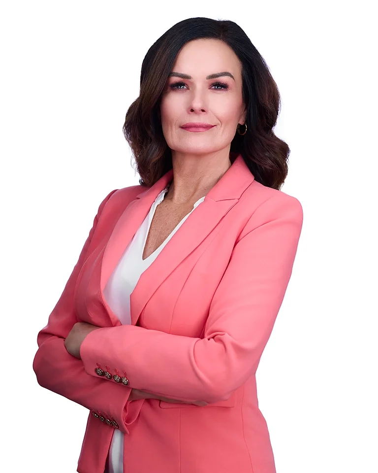 A confident woman with dark brown hair and light skin, wearing a pink blazer and white shirt, standing against a plain white background.