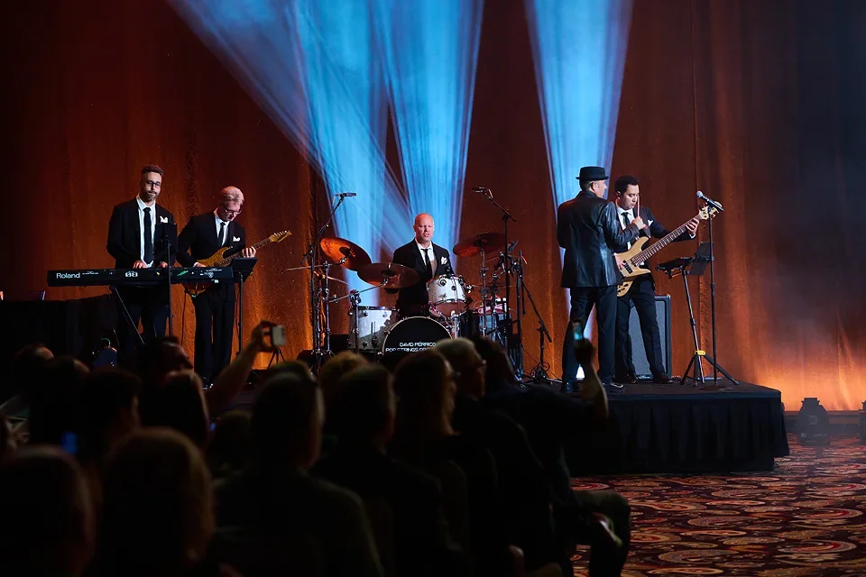 A five-piece band performing on a stage with wood-paneled walls and blue lighting effects, with an audience watching.