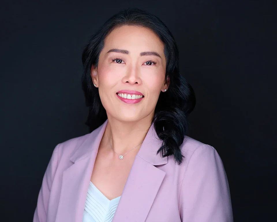 A professional woman with black hair, wearing a purple blazer, white top, and a necklace, smiling against a dark background.