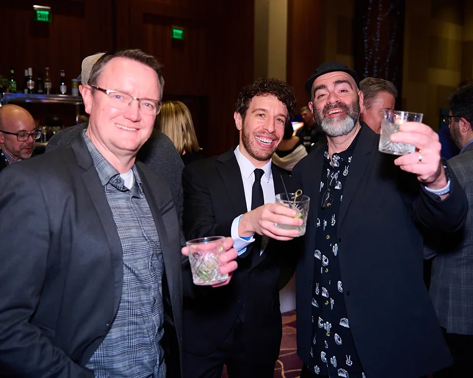 Three men smiling, dressed in suits, holding drinks at a social event or party.