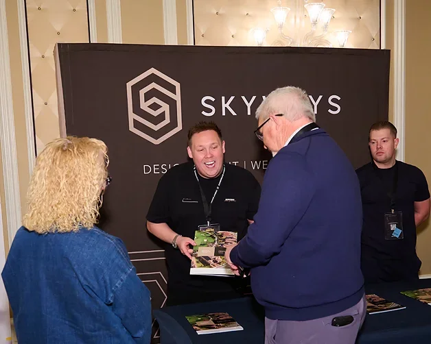 Four people at a booth with a black backdrop displaying the Skyways logo, two men and two women are engaged in conversation, one woman has curly blonde hair, the man on the left is smiling and holding a brochure, and the man on the right is wearing g