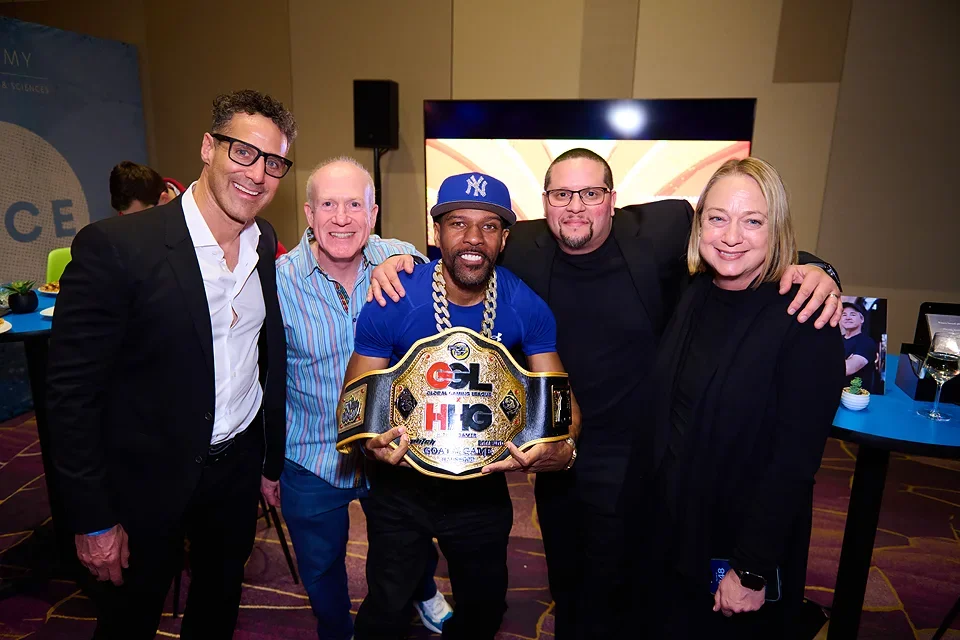 Five people celebrating at an event, with one man holding a championship belt with logos, wearing a blue shirt and cap, and others smiling and posing together.
