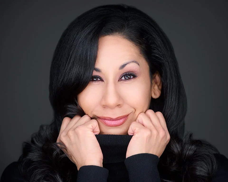 A woman with long black hair, smiling, wearing a black turtleneck, posed with her hands near her face against a dark background.