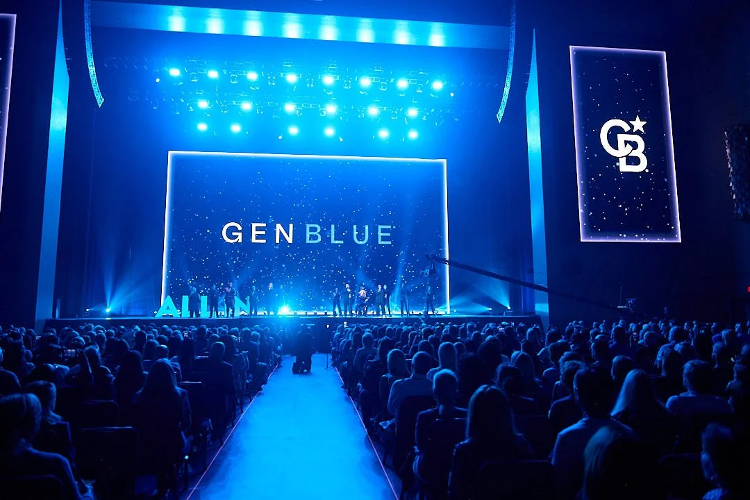 A large audience seated in front of a stage with blue lighting, displaying the words "GENBLUE" on a big screen and to the side, during a presentation or event.