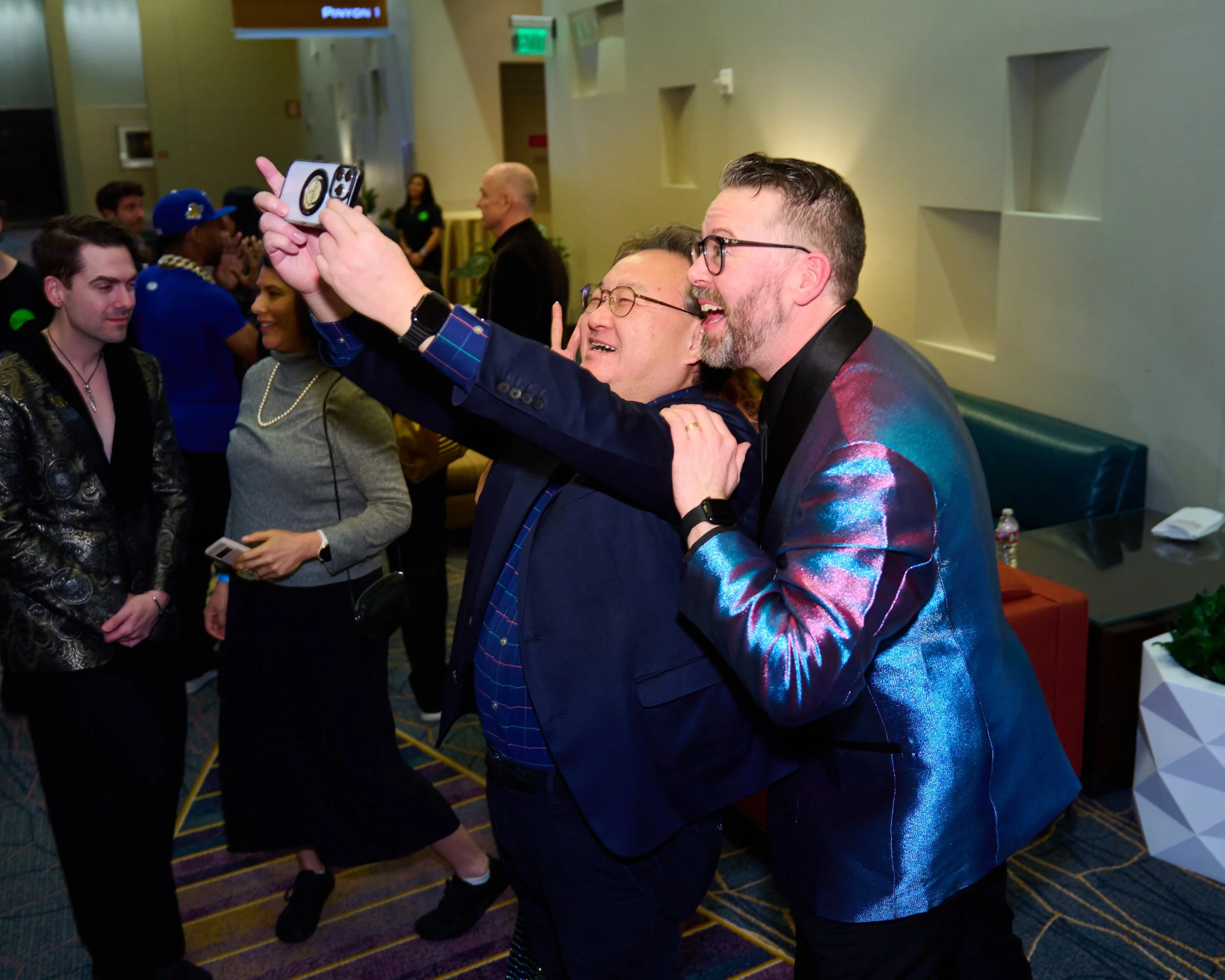 Two men taking a selfie at a party, smiling and embracing each other, with other guests in the background.