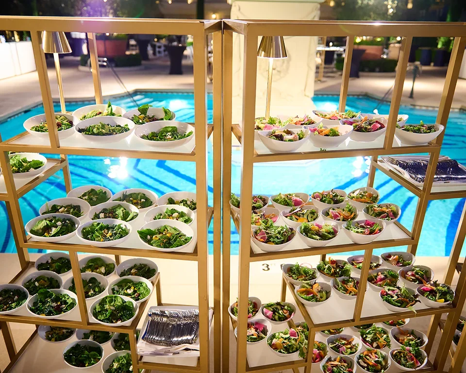 Displays multiple bowls of salad arranged on racks near a swimming pool with bright lighting and outdoor setting.