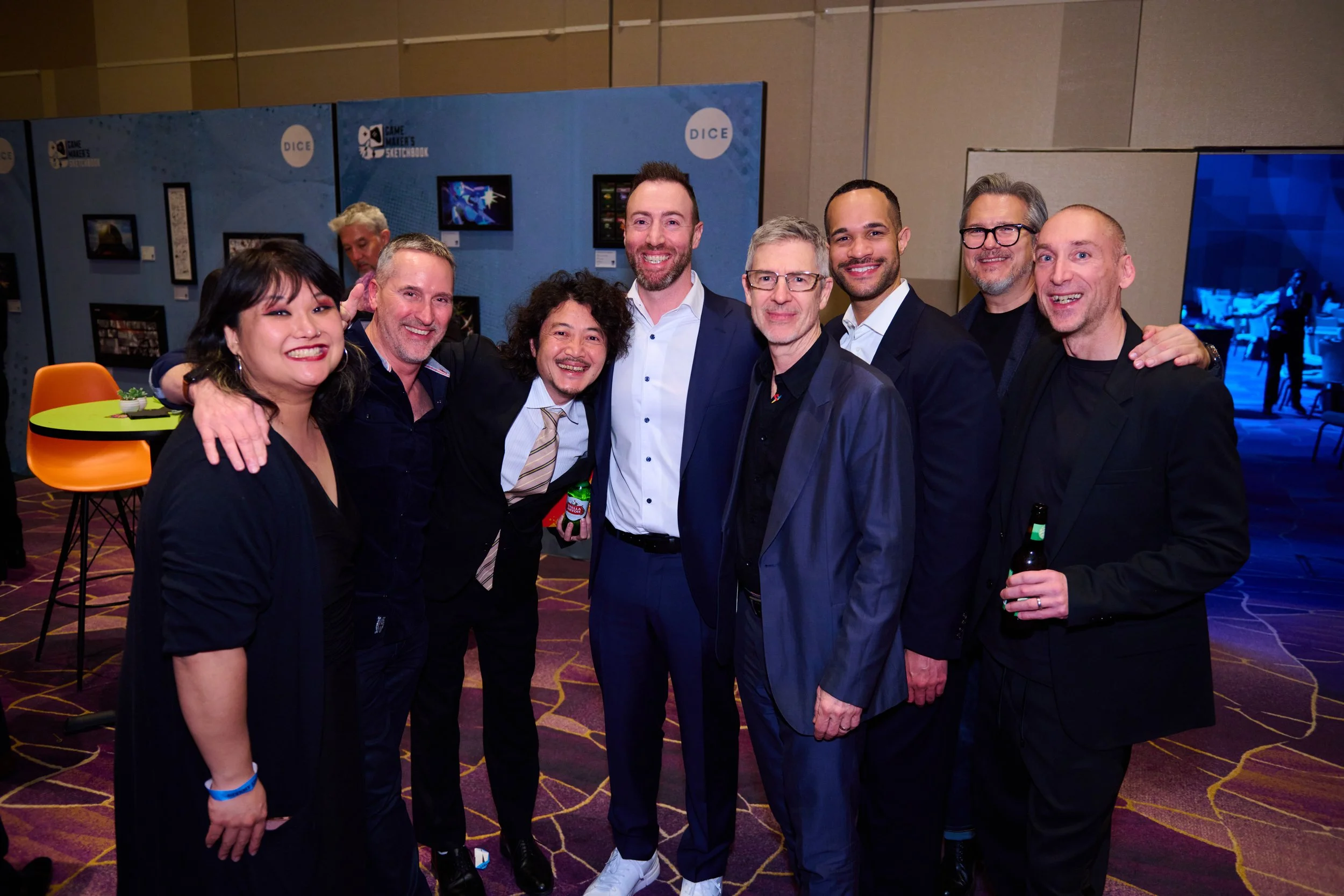 A group of nine people posing together indoors, smiling, with a blue backdrop behind them that has the word 'DICE' and images of screens, suggesting a conference or event setting. Some are holding drinks.