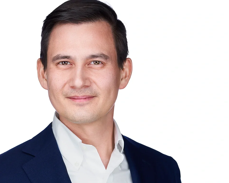 Headshot of a young man with dark hair, wearing a navy blazer and white collared shirt, smiling softly against a white background.