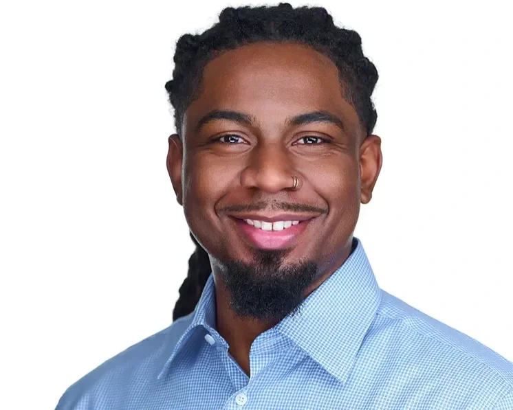 Close-up portrait of a smiling man with dreadlocks, a nose ring, wearing a light blue checkered shirt, against a plain white background.