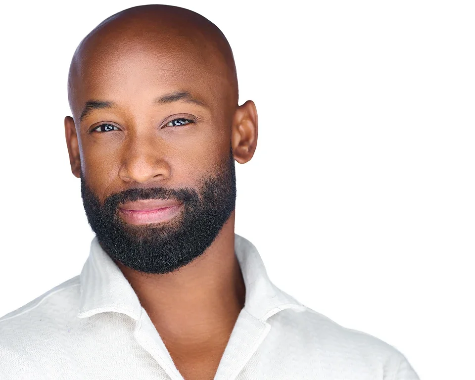 Portrait of a bald African American man with a full beard wearing a white shirt, smiling slightly, against a plain white background.
