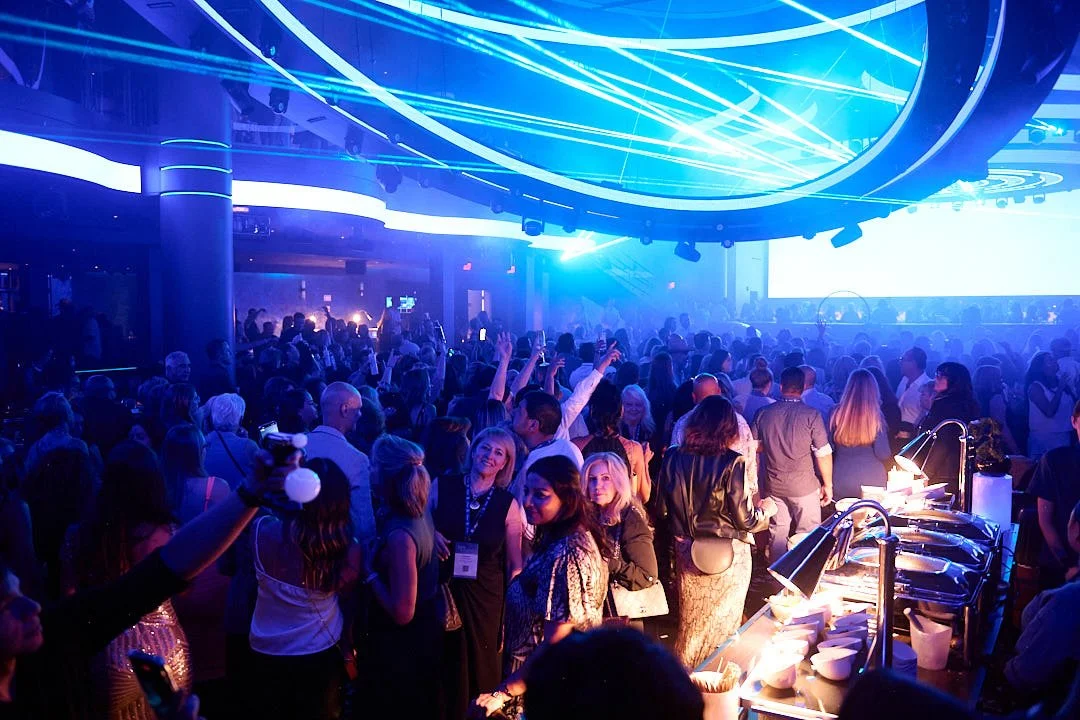 Crowd of people at a nightclub or party with blue neon lighting and a bar area.
