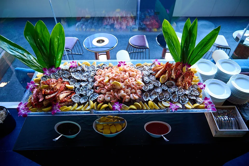 Seafood platter with oysters, shrimp, and lobster garnished with greens and edible flowers, alongside lemon wedges, sauces, and empty plates.