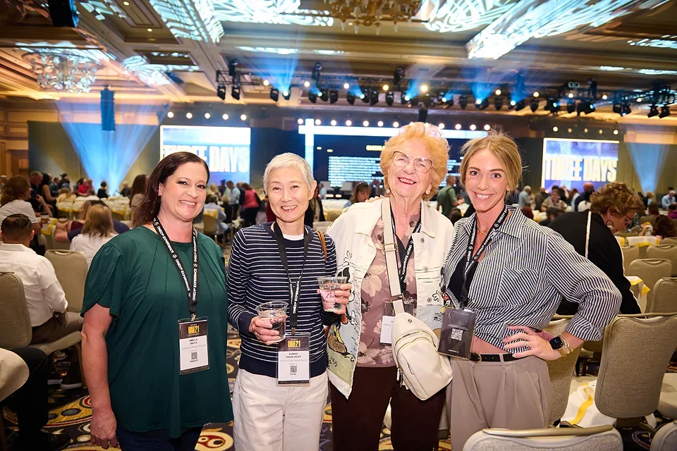 Four women smiling at an event, standing in front of a large stage with blue lighting and a screen that says 'DAYS,' surrounded by seated attendees in a conference hall.