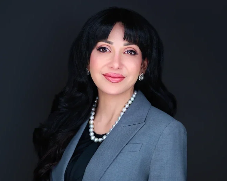 Professional woman with long black hair, wearing a gray blazer, pearl necklace, and earrings, smiling against a dark background.