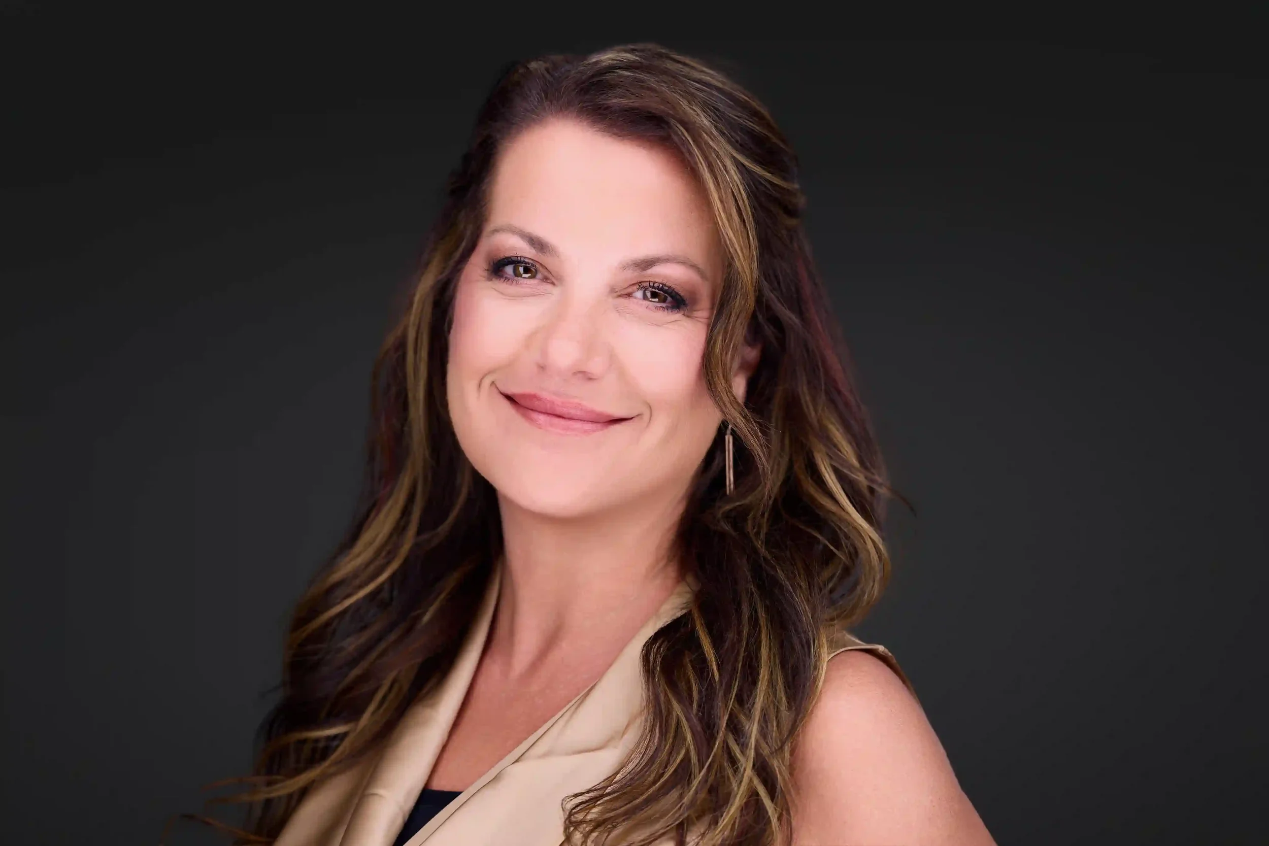 Professional headshot of a woman with long wavy brown hair wearing a beige blazer, smiling warmly against a dark background, photographed at Pandorica Headshot Studio in Las Vegas