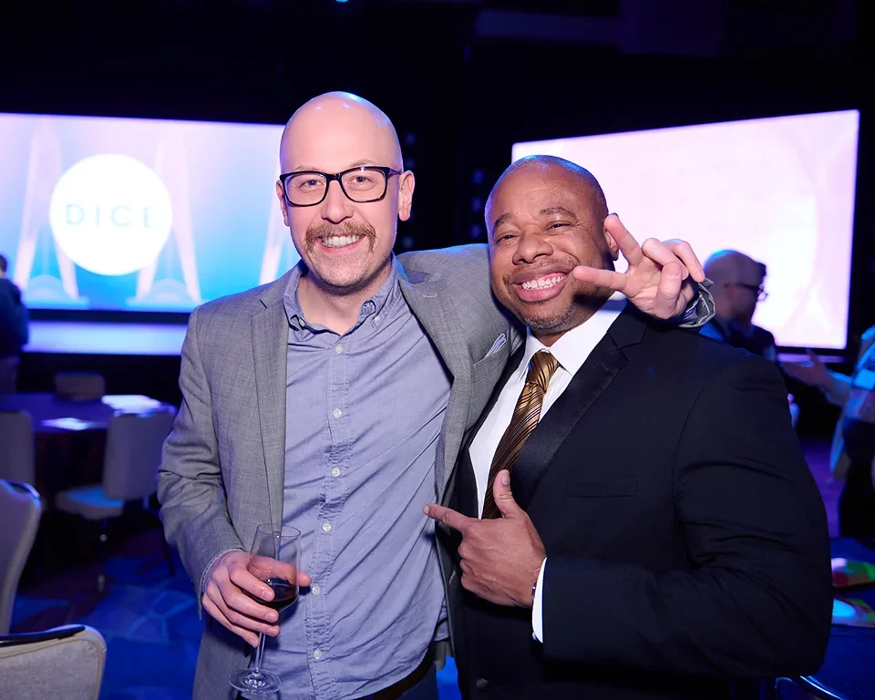 Two men in suits smiling and posing for a photo at an event with a large screen displaying 'DICE' in the background.