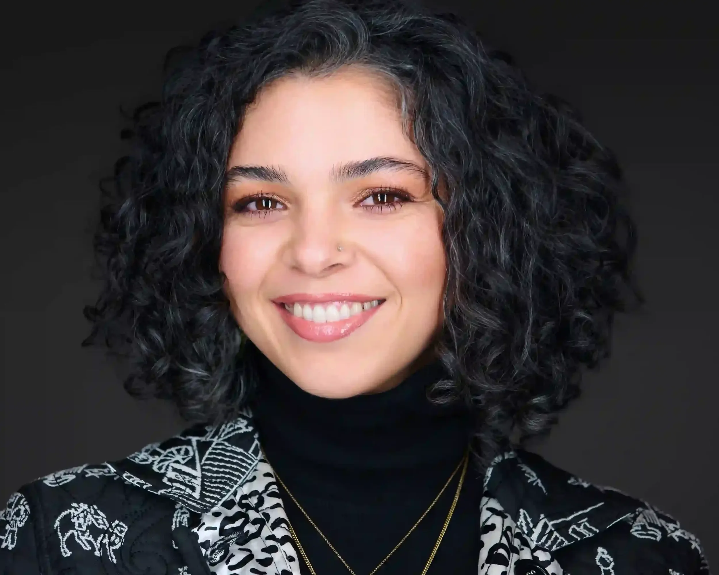 A young woman with curly black hair, wearing a black turtleneck, a patterned blazer, and gold jewelry, smiling against a dark background.