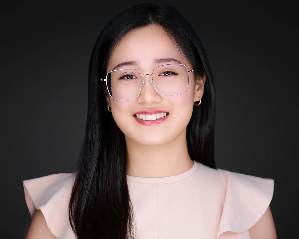 A young woman with long black hair, wearing glasses, earrings, and a light pink top, smiling against a dark background.