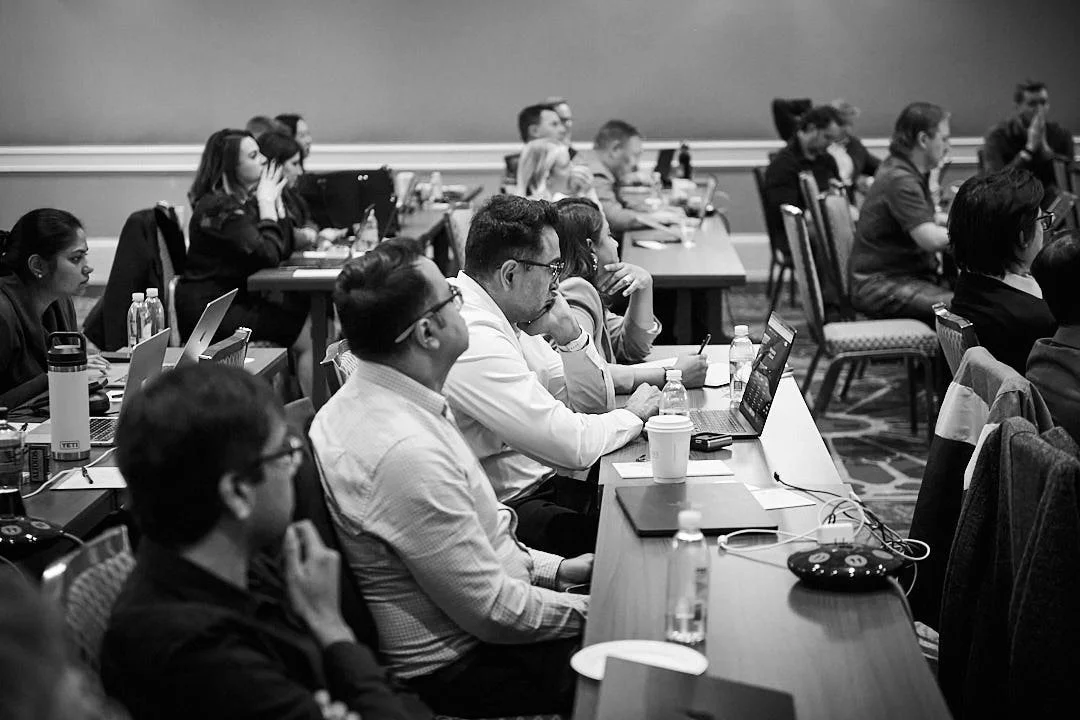 People attending a conference sitting at long tables with laptops and water bottles, listening attentively.