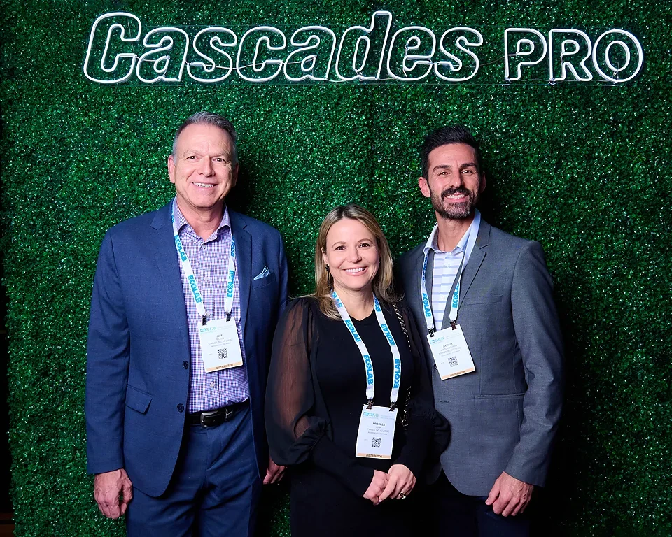 Three people in business attire smiling at a conference with a green leafy backdrop and a neon sign that says 'Cascade PRO'