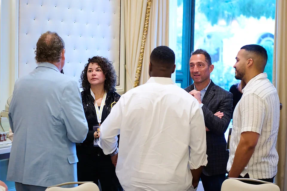 A group of five people engaged in conversation at a professional or social event inside a decorated room with large windows and curtains.