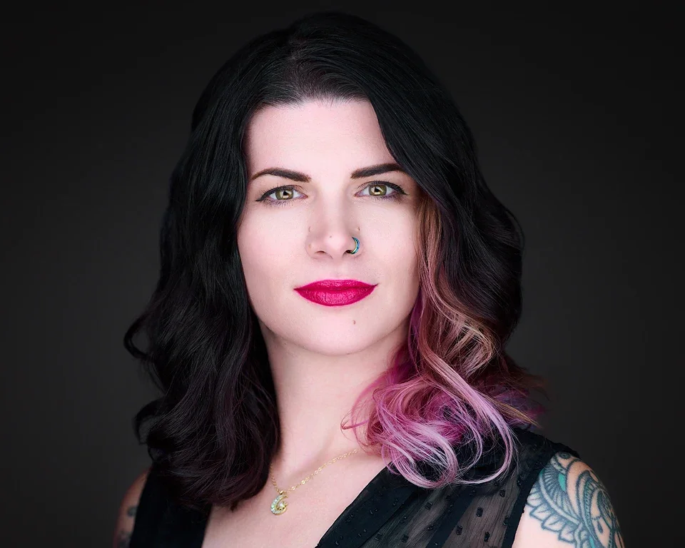 Portrait of a woman with dark wavy hair with pink highlights, wearing a nose ring, bright lipstick, and a necklace, against a dark background.