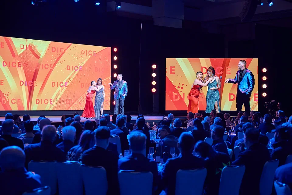 Four people on stage performing at an event with an audience seated in front of them, large screens behind displaying the word 'DICE' with colorful graphics.