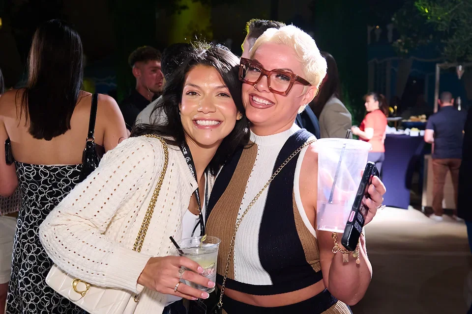 Two women smiling and hugging at a social event, one holding a drink and the other with a large cup, with other people in the background.