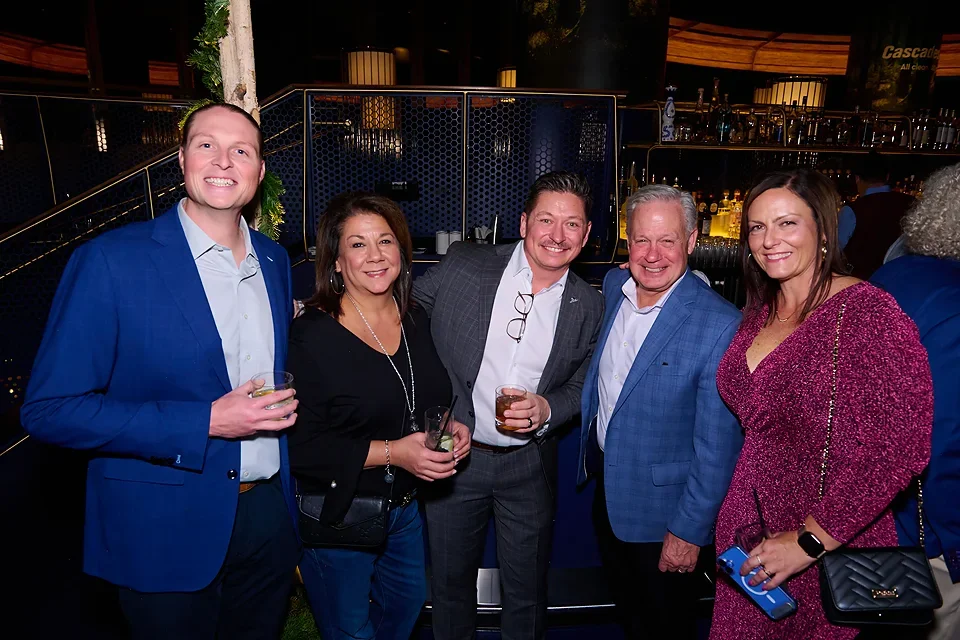 Group of five adults smiling at a social event, holding drinks, with bar and dim lighting in the background.
