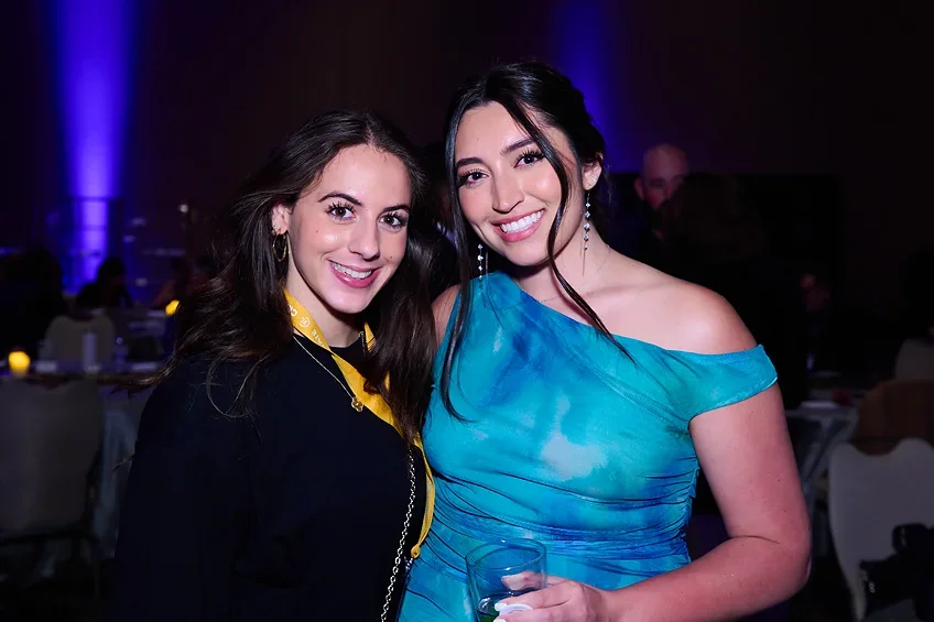 Two young women smiling at a party, one holding a drink, wearing a blue dress, and the other in a dark top with a yellow lanyard, in a dimly lit room with tables and people in the background.