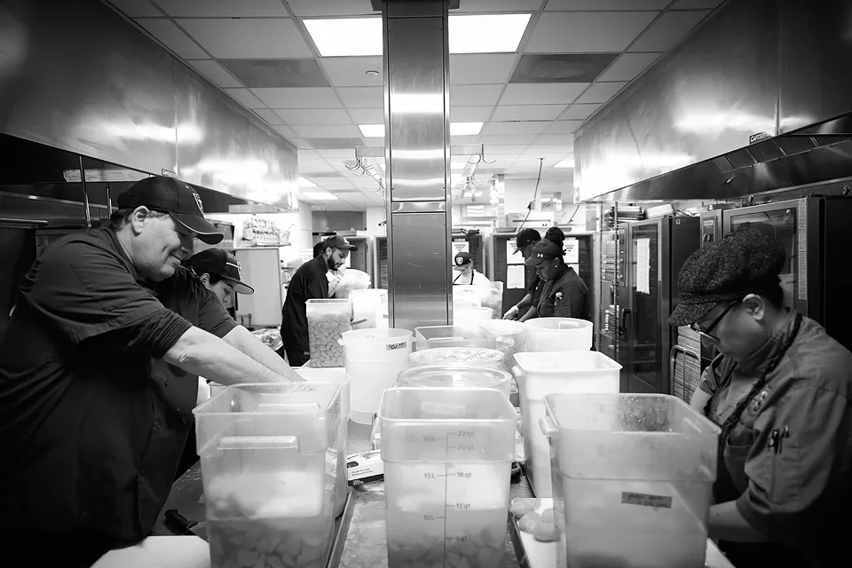 Kitchen staff working at a counter with large plastic containers in a commercial kitchen.