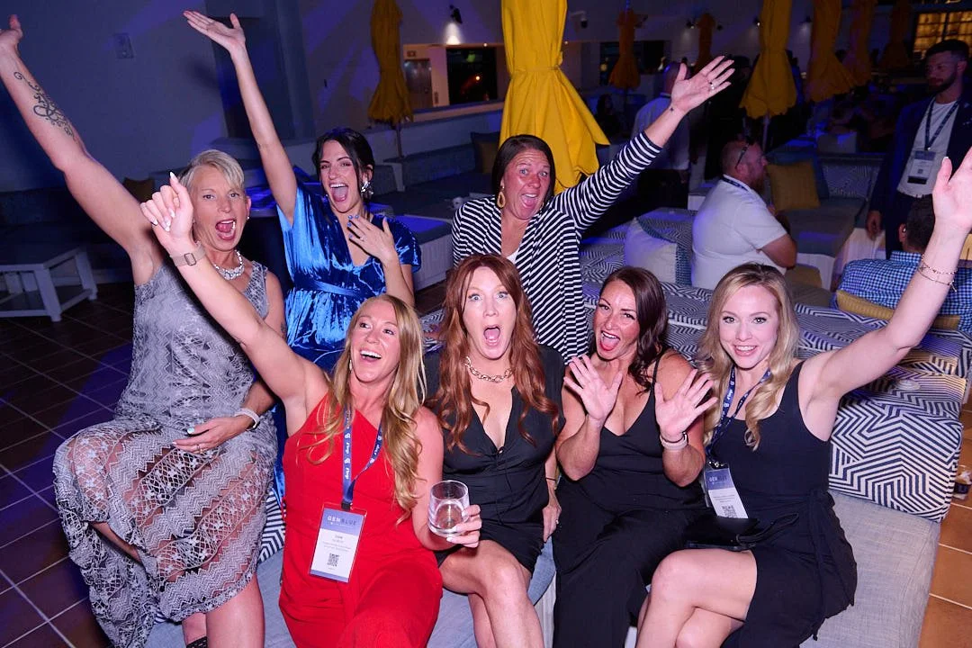 Group of eight women celebrating at a party or conference, with some women sitting and others standing, all smiling and raising their hands in excitement.