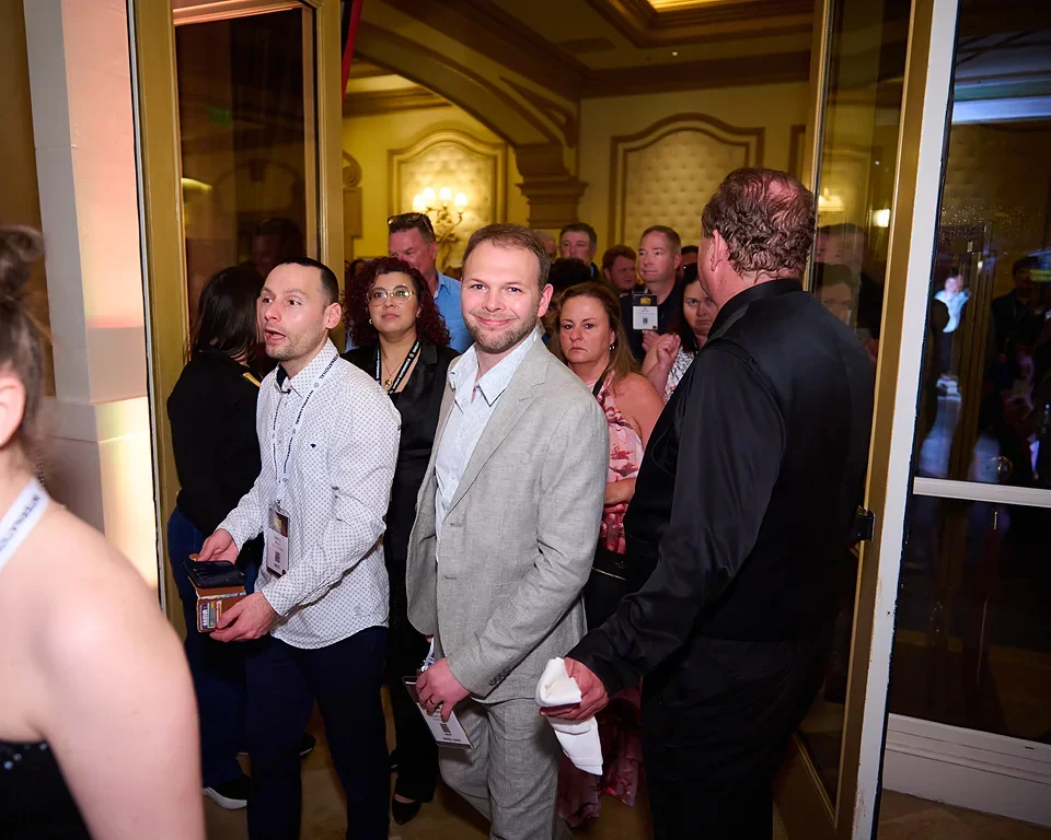 People at a crowded indoor event, lined up at a doorway, some holding badges or tickets, with a man in a gray suit smiling at the camera.