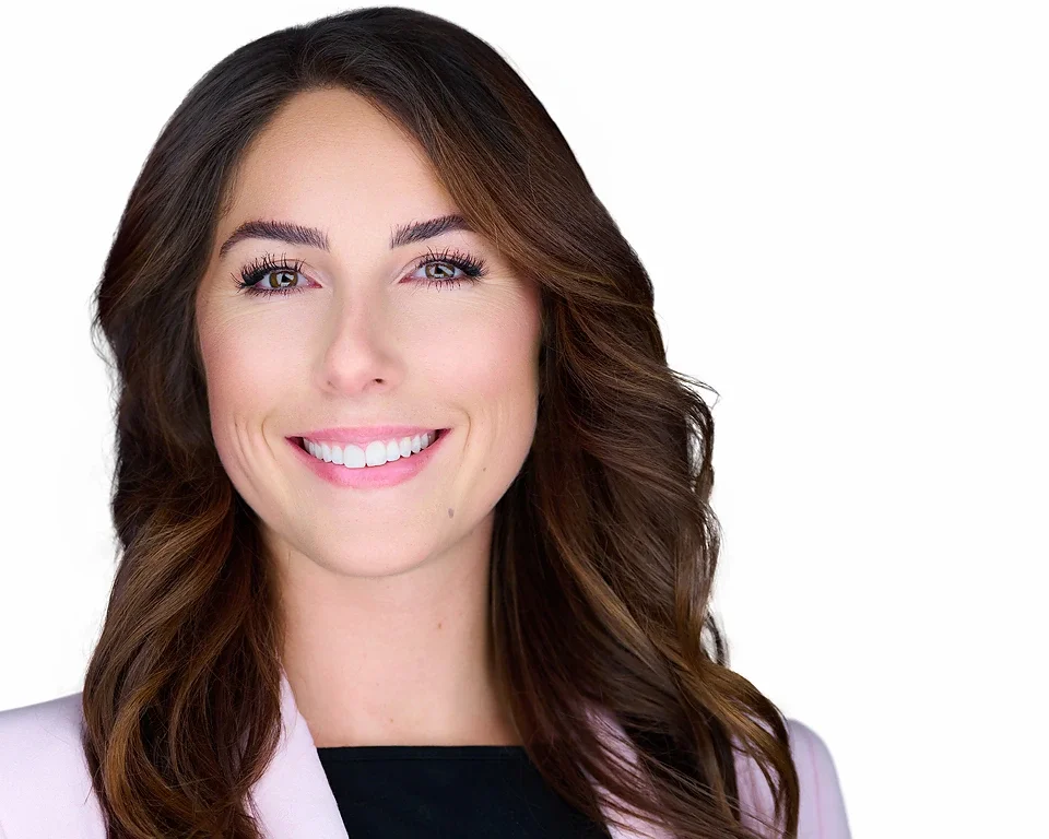 Close-up portrait of smiling woman with wavy brown hair, wearing a light pink blazer and black top, against a white background.