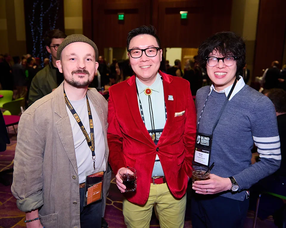 Three men at a social event, smiling and holding drinks, with a crowded room and festive decorations in the background.