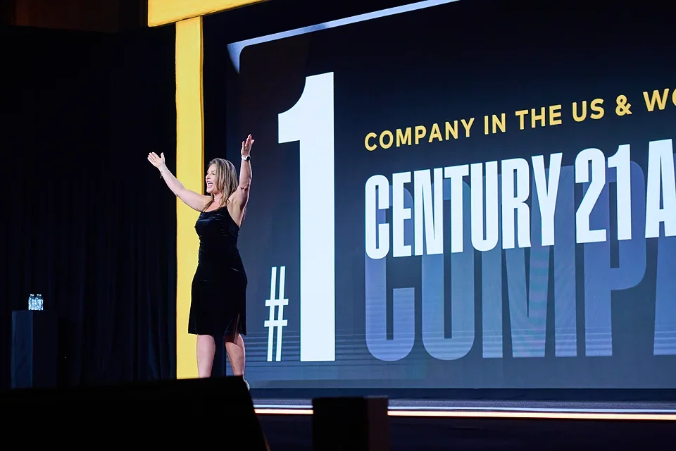 Woman in black dress on stage with large screen behind her displaying '#1 Century 21' and part of a phrase about being a top company in the US and worldwide.