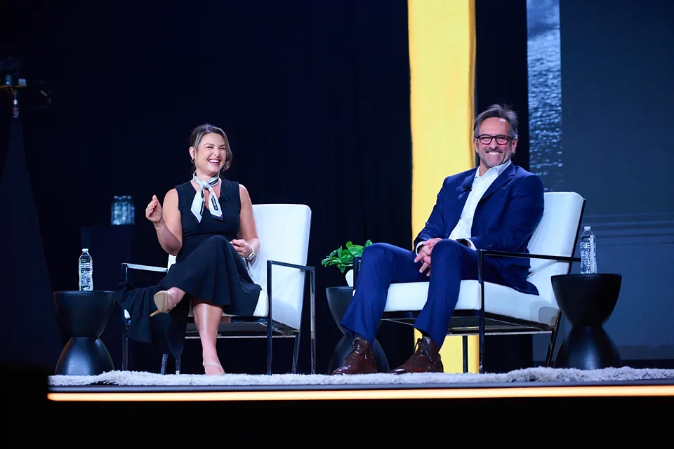 A man and woman sitting on stage chairs, smiling and talking during a panel discussion or interview at an event, with water bottles on small tables beside them.