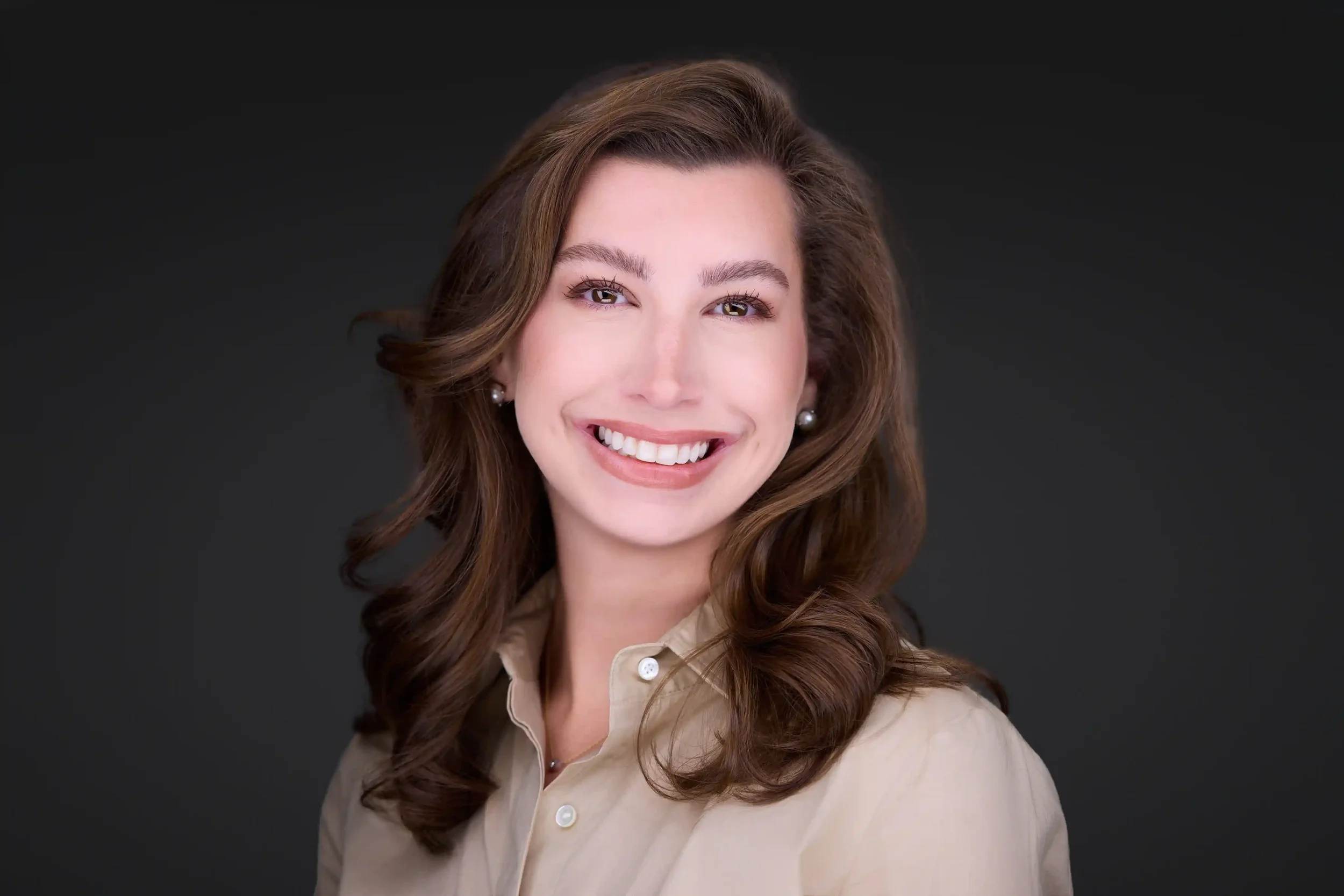 Smiling woman with long wavy brown hair, wearing pearl earrings and a beige button-up shirt, against a dark background.