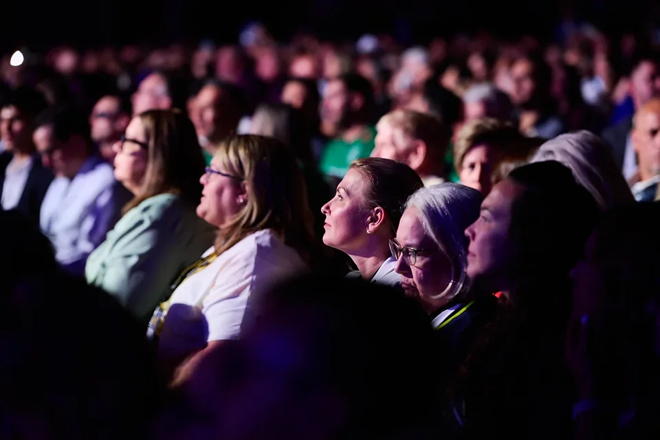 Audience members attentively watching a presentation on stage at a conference or event, illuminated by stage lighting.