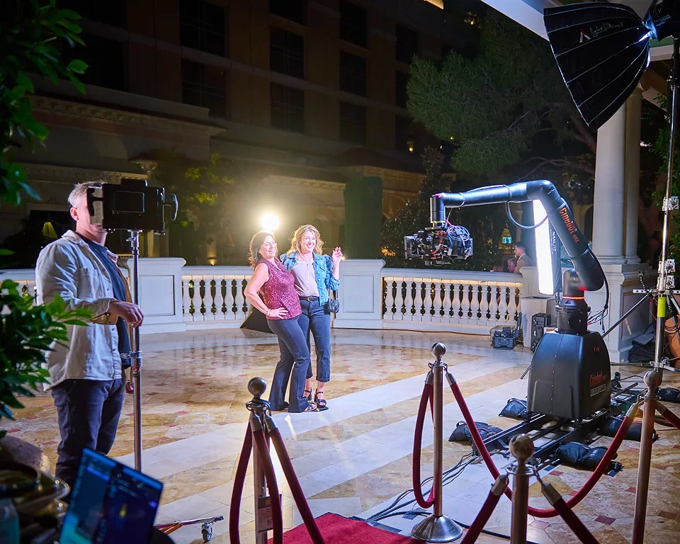 Two women posing for a photo in front of a large camera robot at night, with a photographer and a camera operator nearby, on a marble floor with a white fence in the background.