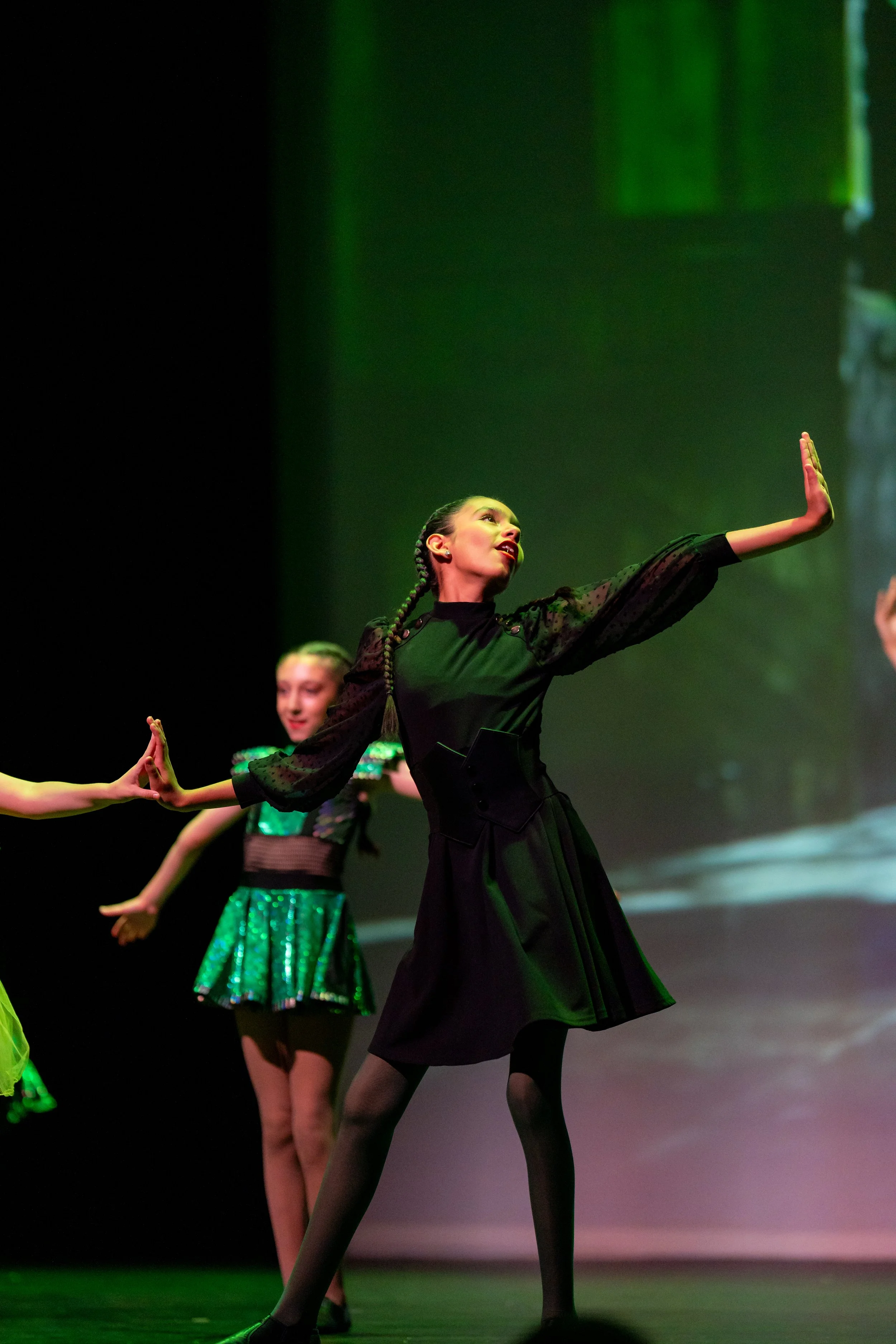 A young girl performing musical on stage, reaching forward with one arm, dressed in a black dress with sheer sleeves, and wearing tights. Other dancers in green costumes are visible in the background, with a stage projection of a scene behind them.